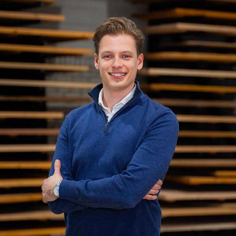 Jonge man met gekruiste armen glimlacht voor gestapelde houten planken in Duumaa-werkplaats, passend bij maatwerk tafels en interieur.