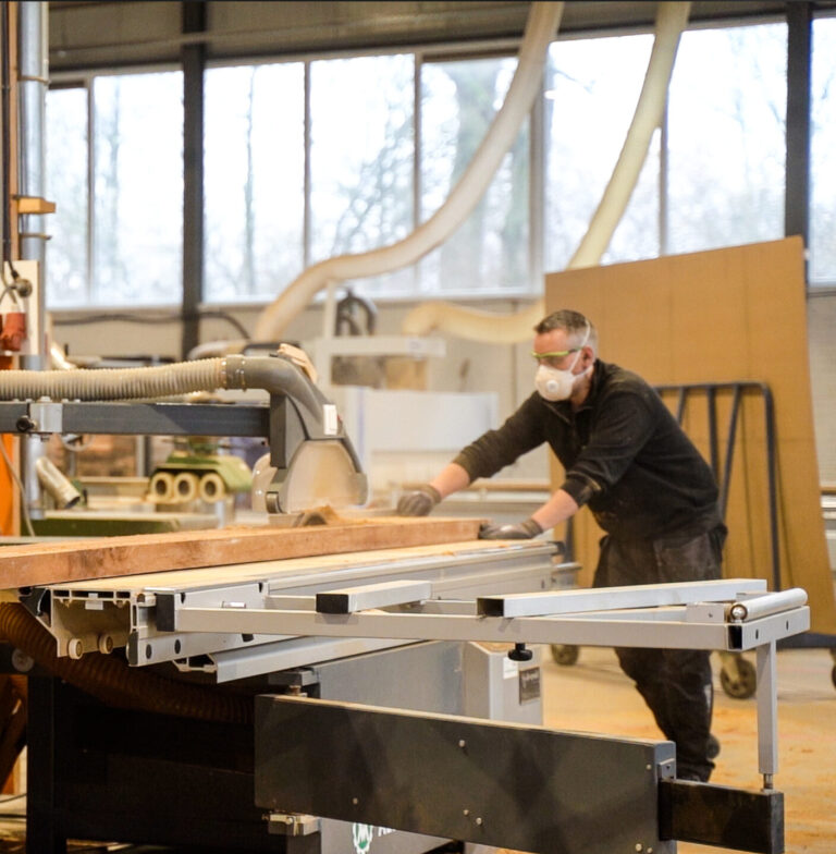 Twee meubelmakers zagen en verplaatsen een lang houten tafelblad voor een maatwerk tafel in de eigen werkplaats van Duumaa. Tekst op jas: "BuitenDuurzaam".