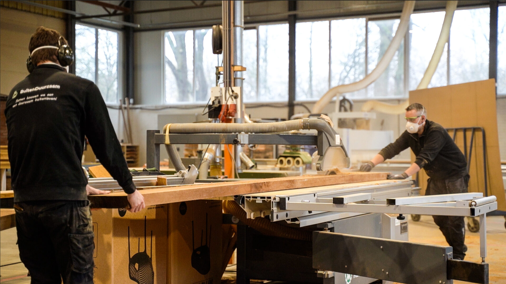 Twee meubelmakers zagen en verplaatsen een lang houten tafelblad voor een maatwerk tafel in de eigen werkplaats van Duumaa. Tekst op jas: "BuitenDuurzaam".